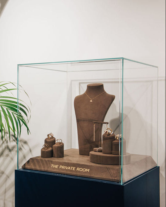A glass display case on a dark pedestal showcasing The Private Room Fine Jewelry, featuring 18K gold necklace and diamond rings.