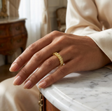 **A RADIANT LEGACY CAPTURED IN GOLD** — ring lifestyle shot, Elegant hand resting on a marble surface, ring on the ring finger