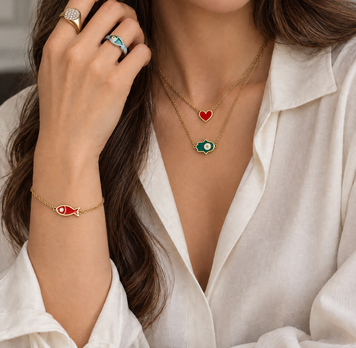 A close-up lifestyle shot of a woman wearing a matching enamel 18K gold jewelry set, including a fish pendant bracelet, a heart pendant necklace, and an evil eye pendant necklace from The Private Room Fine Jewelry