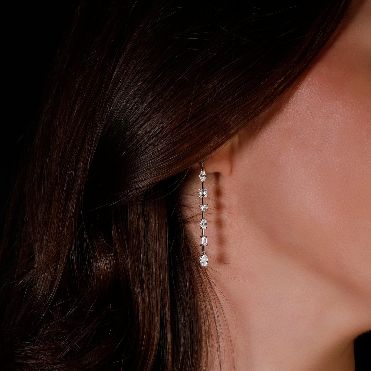 A close-up of a woman's ear wearing a long, delicate drop earring with multiple diamond shapes set in white gold metal, against dark brown hair and a black background - lifestyle