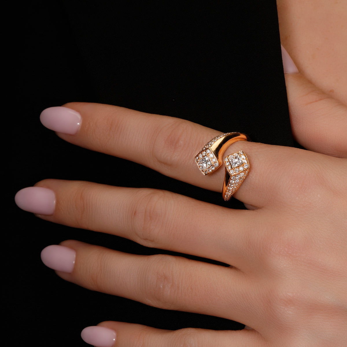 A close-up of a hand wearing a rose gold wrap ring with pavé-set clear stones, set against a solid black background.